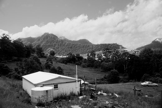 StoddardsChRd-house-mono Looking north over undulating farmland to not-so-distant ranges. The setting was idyllic. Construction of the verandah and awning was underway at the house when this image was made. Poles harvested from the nearby forest have been placed in the ground ready for roofing. The house water supply relied on the rainfall stored in the water tank. This was rural living, basic, but enjoyable.