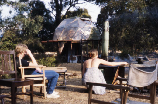 fi-kempsey Cooking on the outside fuel stove at Mungay Creek while Fiona takes it easy. Behind is the geodesic dome, at that stage still clad in plastic. Later, a room was added to the right hand side of the structure and the adjoining downstairs room that contained the kitchen enclosed in log offcuts.
